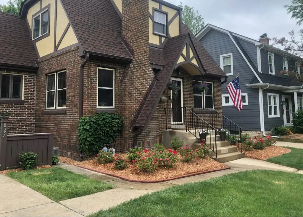 Elegant front yard landscaping of a Tudor-style house with vibrant red flowers, neatly trimmed bushes, and a lush green lawn, adorned with an American flag.