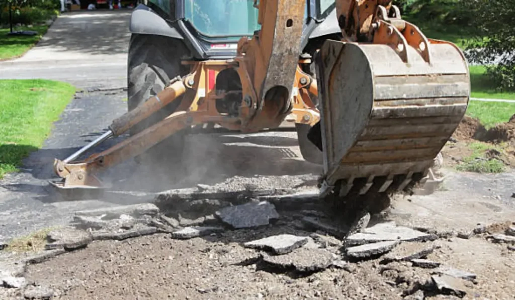 Excavator breaking a cracked driveway into asphalt chunks during demolition at a residential property.