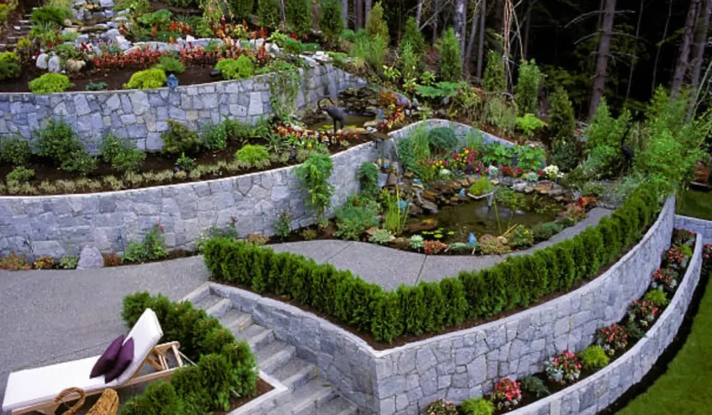 Stone retaining walls supporting tiered garden beds with green shrubs, colorful flowers, water feature, and seating area.
