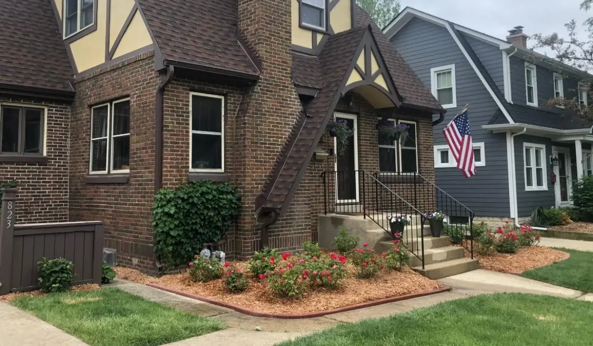 Elegant front yard landscaping of a Tudor-style house with vibrant red flowers, neatly trimmed bushes, and a lush green lawn, adorned with an American flag.